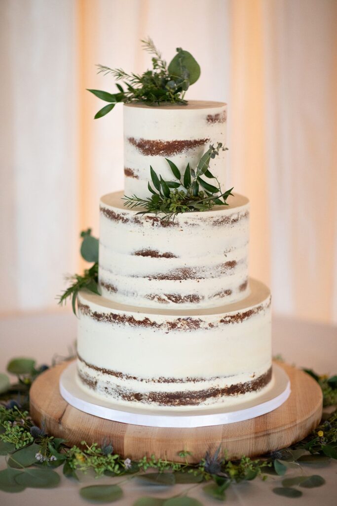 three-tier wedding cake on wood cake stand and greenery around.