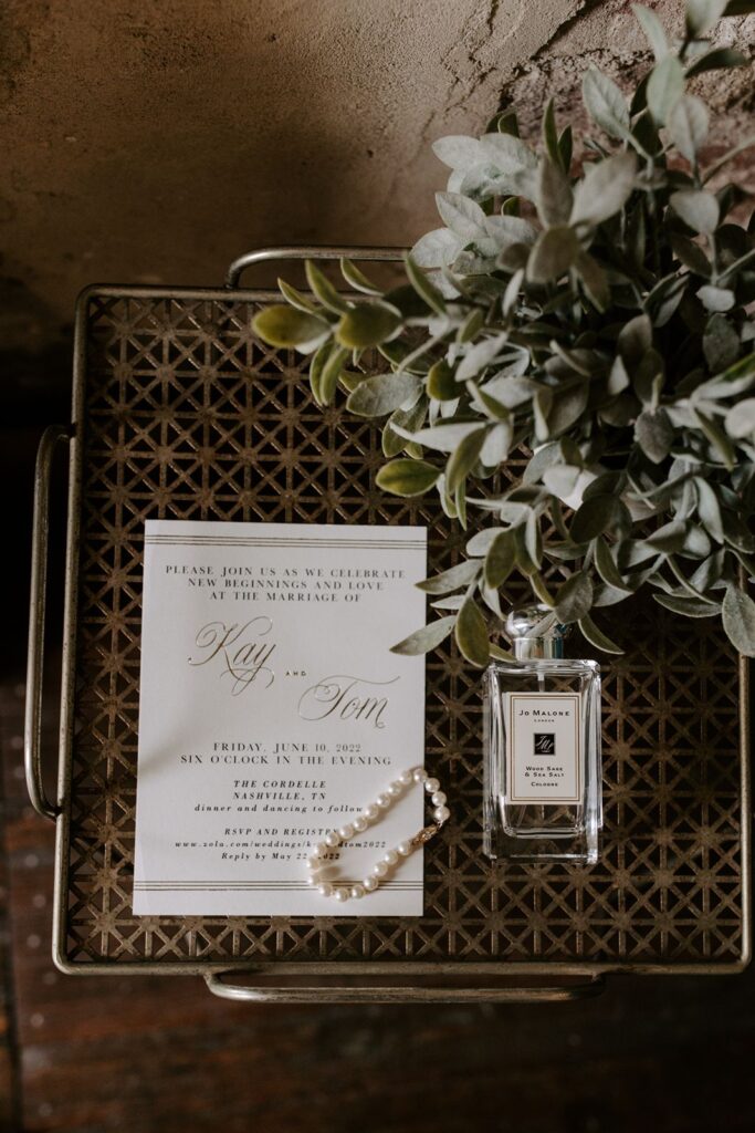 wedding invitation flat lay with perfume, greenery, and a pearl bracelet